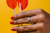 Woman’s hand holding 3 red heart-shaped lollipops like a bouquet, against an orangey-yellow background.