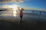 Author’s Mom waving her arms at sunset on a beach.