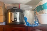 rice cooker, paper products and a plastic container all stored on top of a cabinet