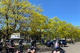 A line of police across the bottom of the photo. Behind them are a row of trees in Lents Park. A police SUV and more officers are visible in the background. Blue sky appears above the scene.