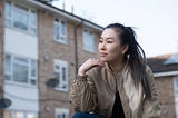 A young person is sitting outside in front of an apartment building. They are Asian and have light skin and long, dark hair tied into a ponytail. They are wearing a brown bomber jacket, and are looking off into the distance with their chin resting on their fingers.