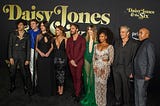 The cast of Daisy Jones and the Six on a black carpet in front of a black and gold background.