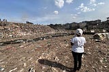 A woman with her back to the camera looks out over a dump site located near several buildings.
