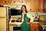 Woman wearing a long green skirt, and matching green vest with a white blouse underneath. She’s in a seventies kitchen with harvest gold appliances and wood cupboards.