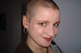 A young woman with a shaved head smiles cheekily at the camera