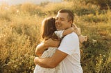 A father hugs his school-aged daughter, smiles, and closes his eyes. The daughter holds a stuffed animal in one hand. The photo is set in a field at sunset, and the plants in the background are illuminated by the setting sun.