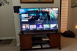 A living room setup with a large flat-screen TV on a wooden stand displaying a movie selection screen. The TV shows the movie Wolfs with the tagline, “What kind of a scam are you running?” Below the main selection, there are smaller options for movies and TV shows like Elementary, Bad Sisters, Lie to Me, and Nature. The TV stand has a dark wood finish with shelves below holding books and an Apple TV box. Small black speakers are positioned on either side.