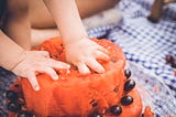 A toddler stocks their hands into a fresh fruit cake made with watermelon and grapes.