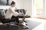 Woman wearing white flowy top and black pants typing on laptop while sitting on black leather chair in living room.
