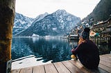 A woman sits on a deck and looks at a lake in winter. She is wearing warm clothes. A mountain in the distance is covered with snow. | Photo courtesy: Riccardo, Pexels