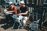 Two people, both wearing hats, are working on typewriters outdoors, both resting on tiny tables. A sign reads ‘Poet for Hire’