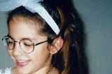 A photo of the author at age four, wearing glasses, a long brunette ponytail, and a white tulle headband.