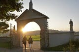 2 people walking hand in hand through an arched gate into the sunset.