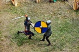 Two men in medieval knight garb fight with swords and shields.