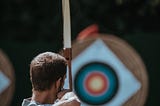 View of a man’s back as he uses a bow to draw an arrow, preparing to fire at a circular target in the distance.