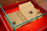 An open book with handwriting and a photo of Anne Frank sits in glass display case on a red platform.