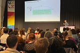A speaker at UXLx presents a slide titled “AI Design Pattern Framework” to a seated audience.