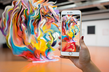 A person’s hand holds a smartphone, capturing a vibrant, abstract art piece on its screen. The artwork, a whirl of vivid colors and organic shapes, fills the exhibition space, contrasting with the minimalist gallery walls.