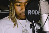 1300Liko in the music studio wearing a white t-shirt with brown colored braid tips resting over his right shoulder. He has on a gold necklace and a diamond studded charm that says, Black Wealth. A rode microphone pop filter is shielding the right side of his face from full view as 1300Liko looks down at his smartphone to see his song lyrics before recording on the platinuum colored condenser microphone in front of him. Photo by 1300Liko via Instagram.
