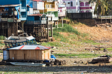 The image shows a riverside community in the Amazon Basin experiencing the effects of severe drought. The riverbed is mostly dry, exposing boats that are stranded on the ground. The houses, some of which are built on stilts, indicate that the area usually experiences high water levels. The dry conditions illustrate the drastic impact of droughts on these communities, affecting their transportation, access to goods, and overall way of life. This visual evidence underscores the findings of the rec