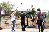 Several production assistants and an audio operator circle a camera operator working on a film/video project at an outdoors location