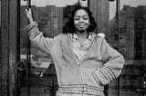 A young Black girl with brown skin and a proud smile stands in front of a Brooklyn brownstone in 1969.