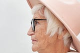 An old woman with short, white hair, a cream hat, and black-framed glasses sits in profile against a white background.