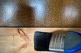 A paint brush beld against some bare skirting board. The background is a dark shiny wooden surface.