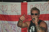 Chris Jericho of rock band, Fozzy, singing on stage in front of a St George’s Cross flag that says “UK Loves Fozzy”.