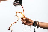 Coffee being poured from a metal jug into a small glass, splashing out over the top and over the hand of the outstretched arm holding the glass.