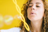 Adult woman blowing bubbles against yellow background.