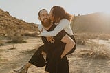 Man carrying his girlfriend on his back, as she kisses him on the cheek, in a desert sunset.