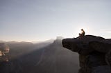 A person sits at the top of a cliff at sunrise