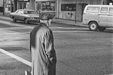 A Vancouver street scene, 1979, and elderly man in a cap and trench coat stands at a corner as cars pass.