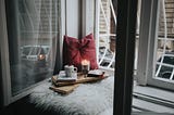 a pillow, candle, mug and books in a corner by a window