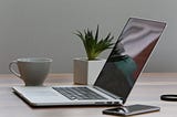 A laptop sits in a desk alongside a coffee cup, cell phone, and a small potted plant.