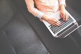 An overhead shot of a femme person in a checkered orange shirt. All that is visible is her white hands with red nailpolish and a ring typing on a Macbook. She is sitting on a grey couch.