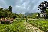 A scenic garden with a flower archway leading into the distant green mountains and semi cloudy blue skies.