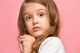 Child with long hair and big eyes. Their hands are folded under their chin, and they look worried or frightened.