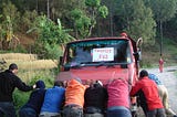 Eight men are pushing the front of a red pickup truck with a sign in the windshield that says “Tourist Bus.” There are trees along the side of the road.