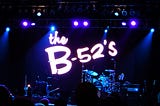 Stage with lit with spotlights and projection of B-52’s logo behind drum kit.