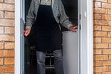 An elderly man in an apron standing at the open external door of his house