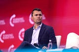 A picture of Charlie Kirk sitting down and turning his head, his face showing a neutral look. He is wearing a black suit with a plaid design, along with a white button-down shirt. He also has a microphone in his hand. The background is red with “Turning Point USA” logos printed on it.