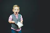 Little boy in collard shirt and vest holding a book with mouth agape in shock at what he read.