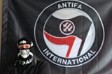 A black flag with the Antifa International emblem, consisting of the traditional antifascist red & black flags with three downward arrows. There is a person standing in front of the flag wearing an FC St. Pauli balaclava.