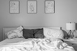 Rumpled cotton sheets on a queen sized bed with three different pillows at the head. Monochrome palette, gray and white.