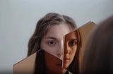 A young woman looks at herself in a broken mirror. Her reflection is split across the shards, showing different parts of her face.
