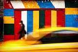 Man in black jacket walking beside red, blue and yellow building