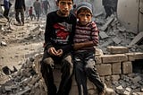 Two children huddle together on a pile of bricks. Famished and fatigued, their fear is palpable. Piles of rubble, broken floorboards, and the refuse of lives lived litter the photo. The two boys — brothers huddle together