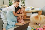 Mom on floor with laptop smiling while boy and girl hug her
